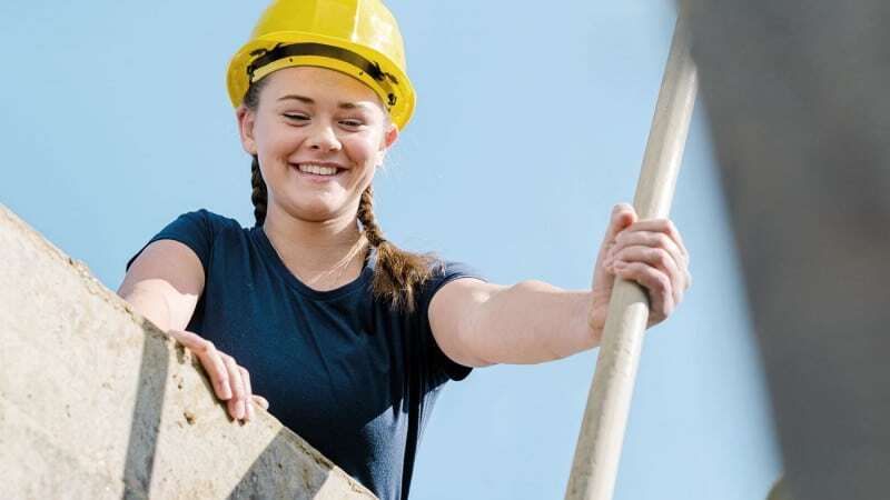 Junge, lachende Frau mit Bauhelm, die von einem Bauwerk herunterblickt